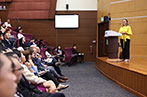 Professor Maxi Scherer on stage delivering her her lecture. She is at a lectern wearing a bright yellow top.