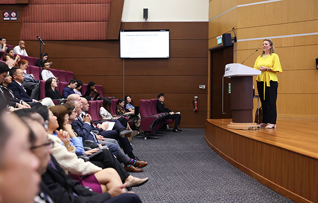 Professor Maxi Scherer on stage delivering her her lecture. She is at a lectern wearing a bright yellow top.