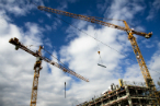 Two yellow construction cranes pictured against a cloudy sky, with the top of an unfinished building in the bottom right-hand corner.