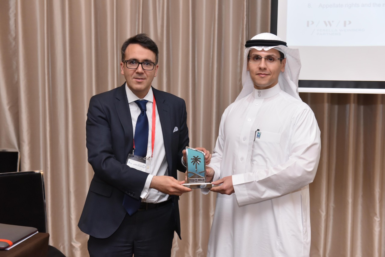 Two men stand together holding an award between them. The man on the left is wearing glasses and a suit. The man on the right is wearing a thwab.