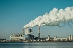 A power station next to a body of water. Smoke is billowing out of the chimneys into a blue sky.