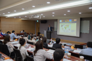 Professor Rafael Leal-Arcas stands behind a podium in front of a lecture hall full of seated students. He gestures towards a screen where his presentation is displayed.