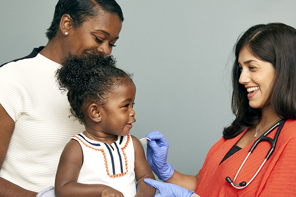 Toddler sitting on parents knee, about to receive a vaccine from a doctor