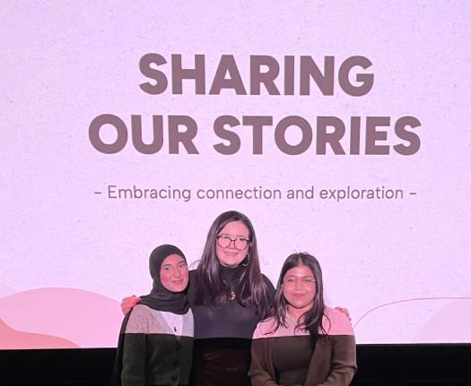 Three female students pose together underneath a banner for their community stories event