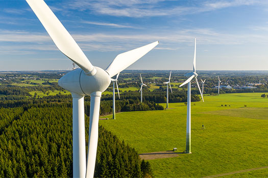 Wind turbines standing in a green field next to a forest on a mostly clear day.