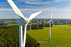 Wind turbines standing in a field next to a forest on a mostly clear day.