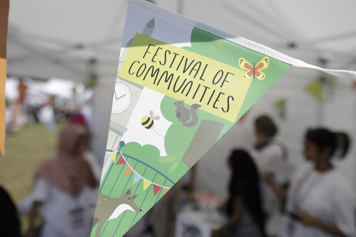 A triangle of Festival of Communities bunting against a blurred background of the inside of a Festival tent. The bunting it white, green and yellow and has pictures of the Queen Mary clock tower, trees, a fox, a butterfly and and a bumble bee on it.