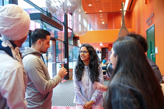 Group of postgraduate students standing and talking in a large open reception with orange and glass walls