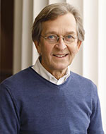 Martti Koskenniemi. He is wearing a blue jumper with a white shirt underneath. He has glasses on and is smiling in front of a pillar.