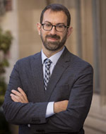 Samuel Moyn. He is wearing a blue checkered blazer and a grey and navy checkered tie. His arms are folded and he is smiling with glasses.