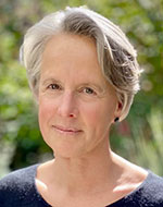 Wendy Brown. She is wearing a navy top, and has short grey hair. She is standing in front of foliage.