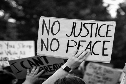 Greyscale image of someone holding up a sign saying 'no justice, no peace' at a protest. Another sign in front says 'I can't breathe'.