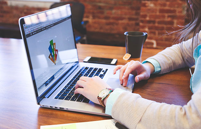 A person sat at a table typing on a laptop. A cup of tea next to them.