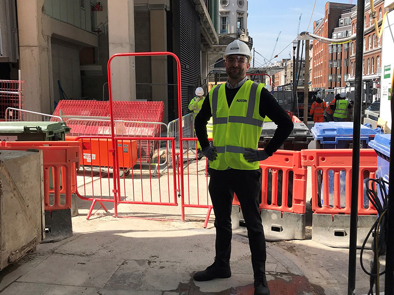 A man in a high vis vest and hard hat standing in a construction site with his hands on his hips and a smile on his face