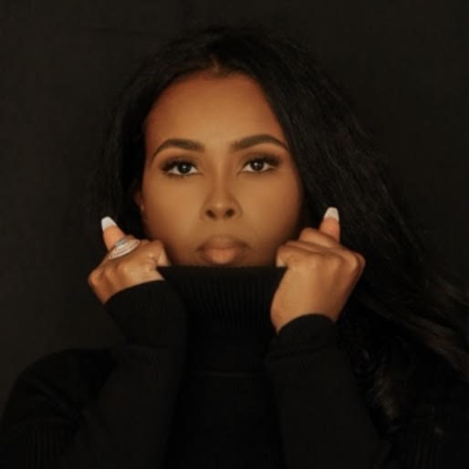 Photo of Hani Garabyare - she is holding the neck of her black rollneck jumper around the edges of her face and the photo has been taken against a black backdrop.