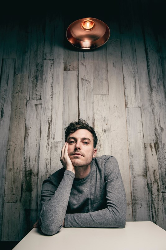 Photo of alumnus, Tom Machell, sat at a table, resting his jaw in his right hand. The photo is taken from a low angle and shows a single light fixture above Tom.