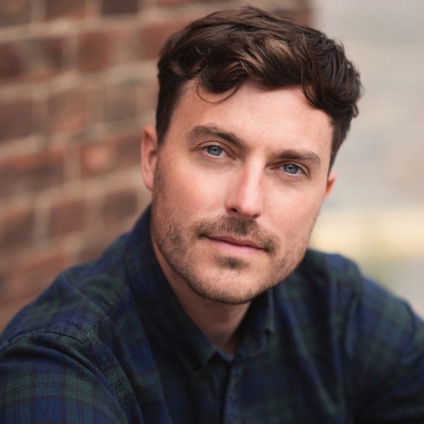 Headshot of alumnus, Tom Machell, wearing a blue and green plaid shirt, sat against a brick wall