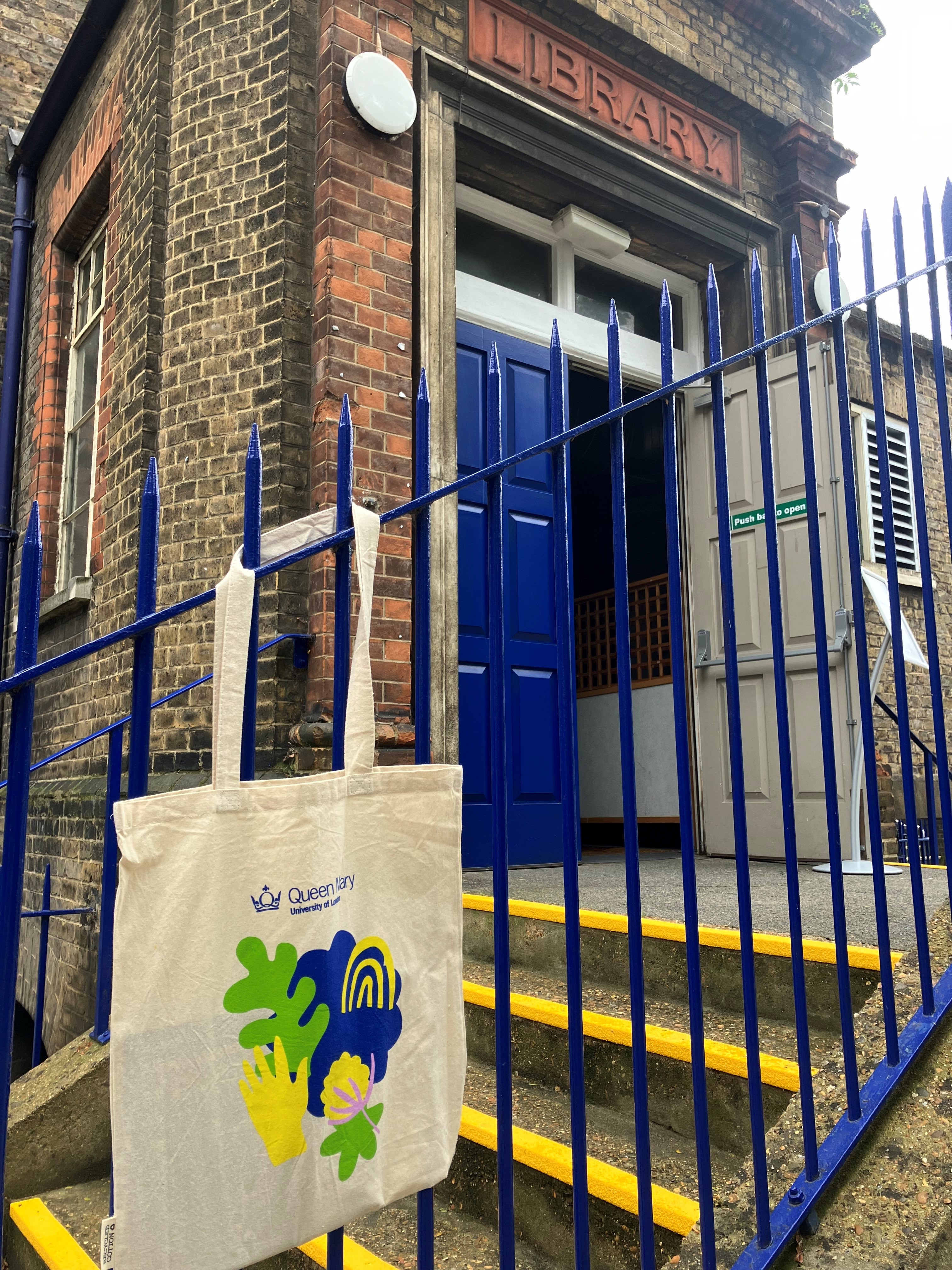 A canvas bag with colourful illustration and Queen Mary logo hanging from a railing in front of the Octagon building