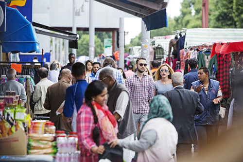 A busy street in east London: in the foreground two women chat by a market stall whilst people walk past in the background.