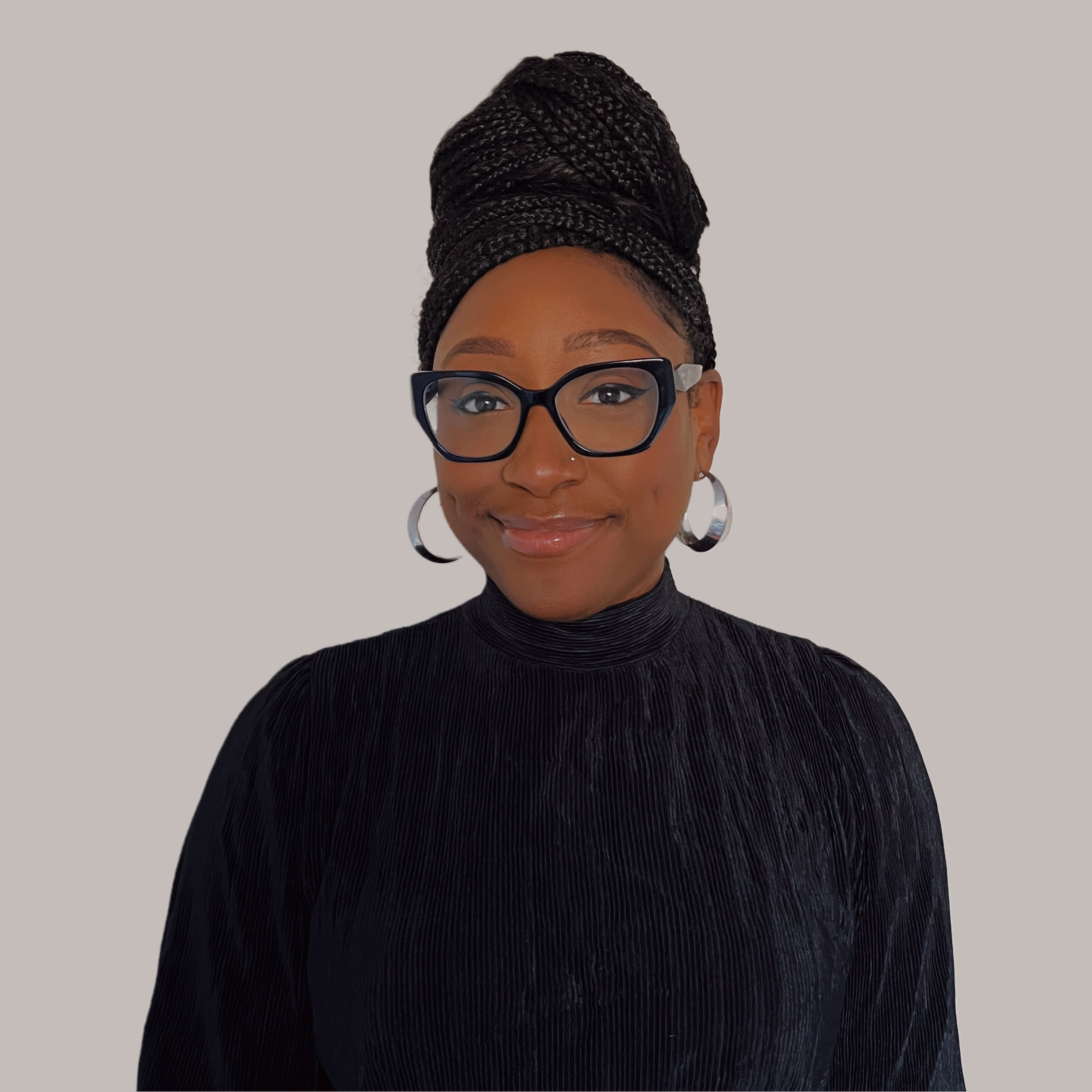 A headshot of Briana: a young Black woman wearing black glasses, a black turtleneck, silver hoop earrings and a silver nose stud. Her hair is braided and piled on top of her head and she is smiling.