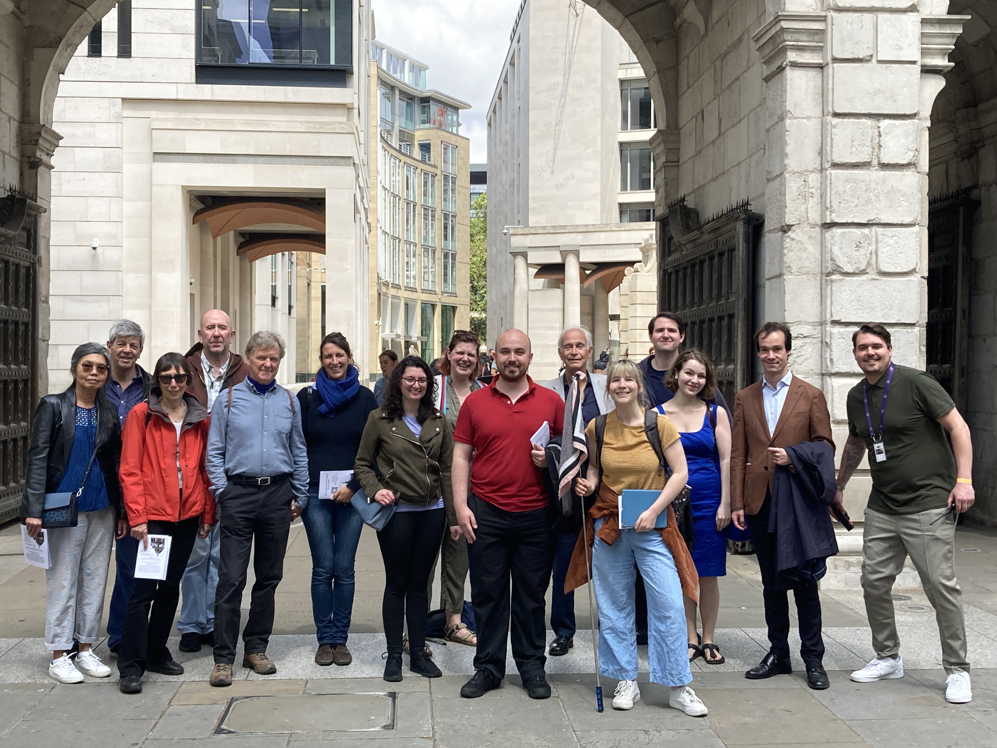 A group of adults dressed for a walking tour with comfortable shoes and layers pose next to Temple Bar after the first tour. Rebecca, a young white woman with blonde hair, stands at the front holding a flag and tour notes.