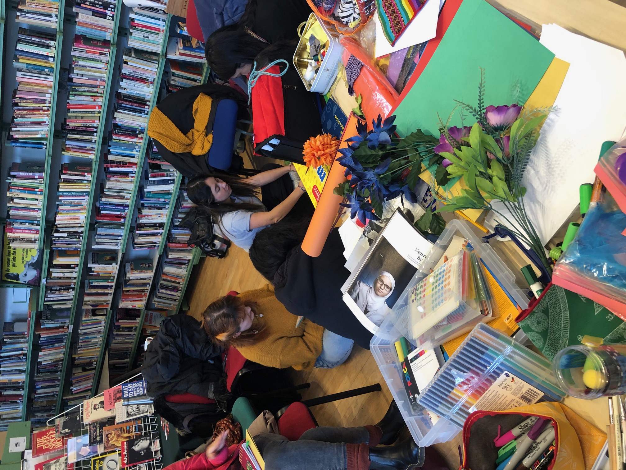 Photo of a busy table covered in zine-making material. In the background people sit on the floor making zines in front of full bookcases.