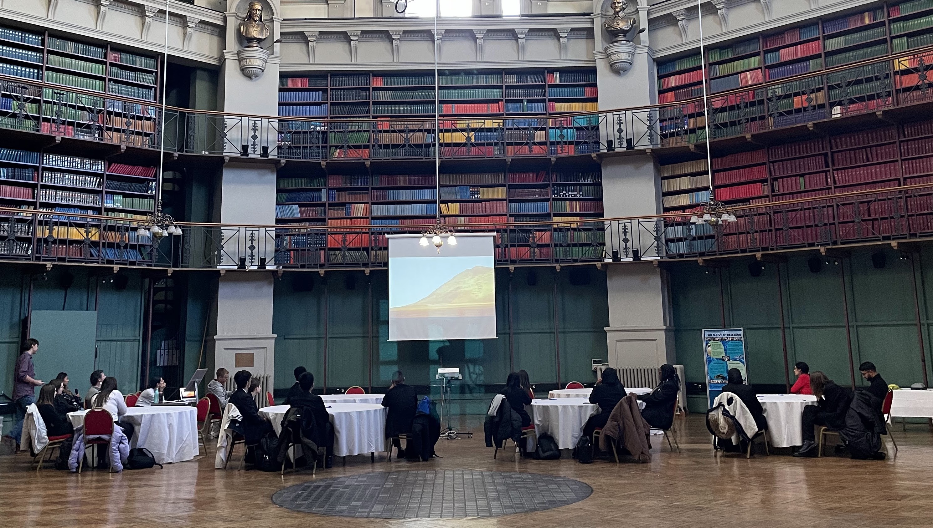 Students sitting in groups at tables watching a live-stream presentation on a projector screen.