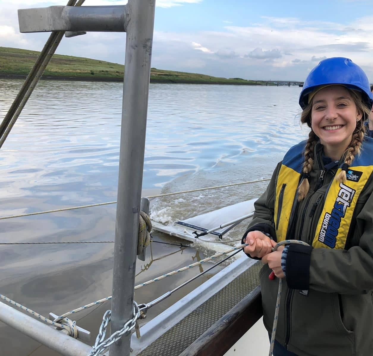 Pauline smiling by the water wearing a hard-hat and a life-jacket.