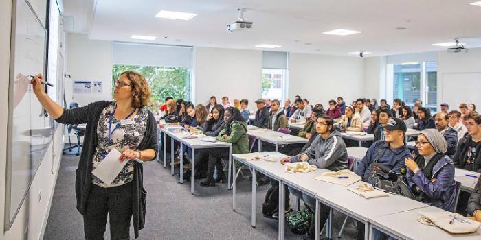 A female lecturer writes on a whiteboard in a full seminar room