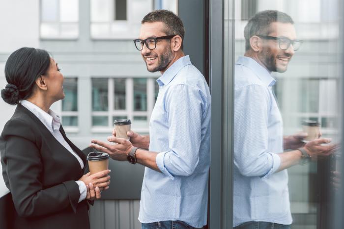 Two people speaking to each other, whilst on a coffeebreak. They are on balcony or terrace and one person's image is reflected in the glass