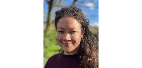 Dr Sarah Milton smiling. She has long curly hair and is wearing an aubergine coloured jumper. In the background is a tree and blue sky out of focus.