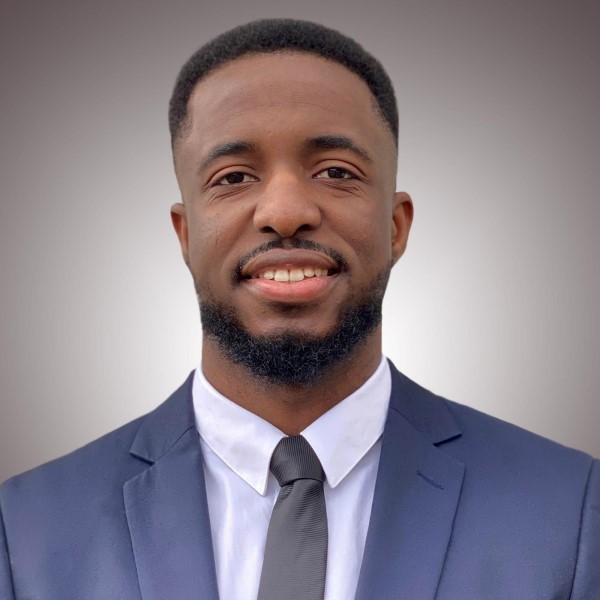 Professional headshot of alumnus, Nicholas Omale. He is wearing a navy suit, white shirt and purple tie.