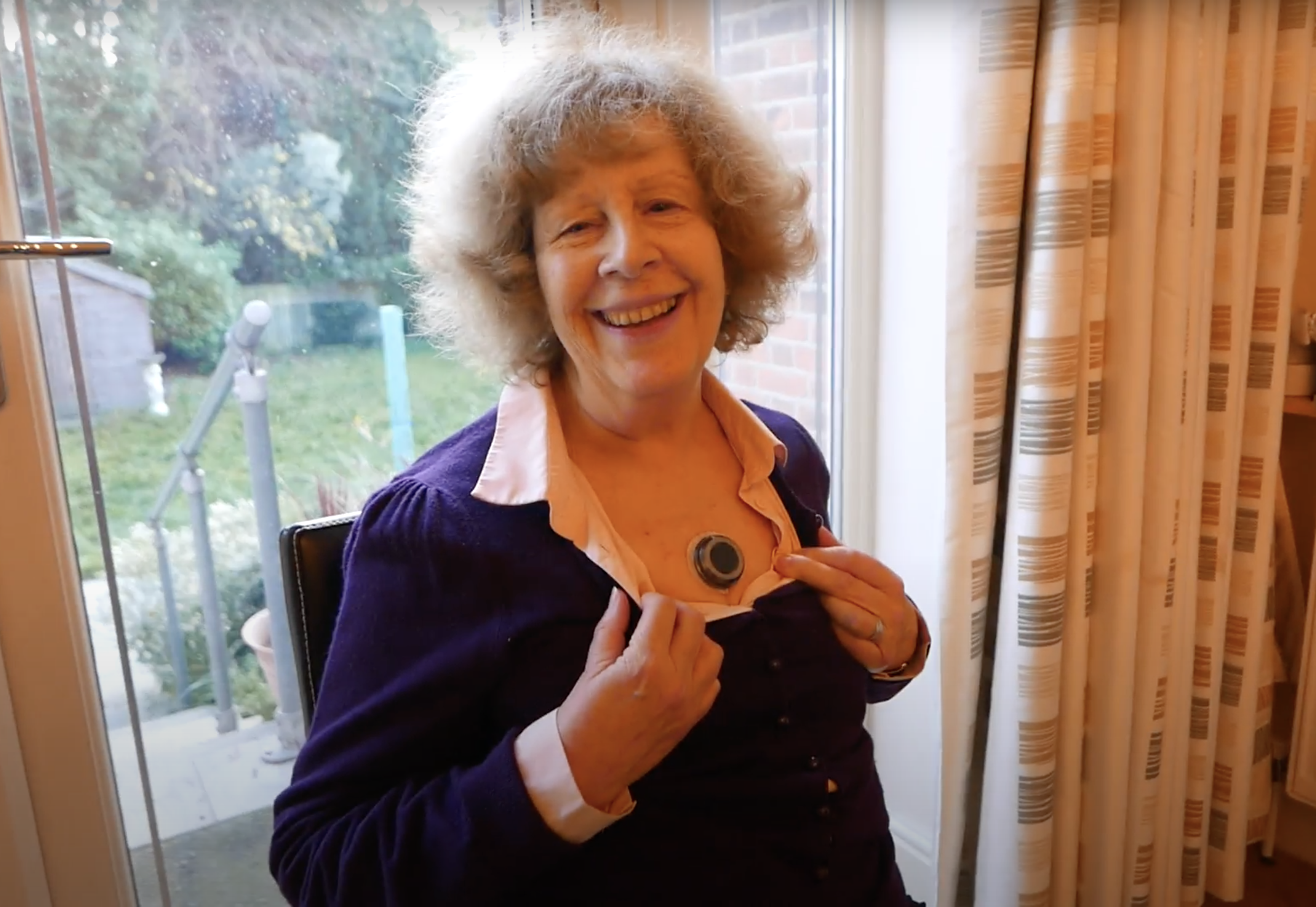 A smiling older woman pulls open the top of her shirt to show the small, black CUE1 device on her sternum.