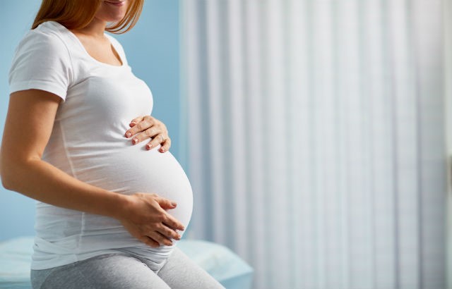 A pregnant woman cradles her bump, while sitting on a hospital bed