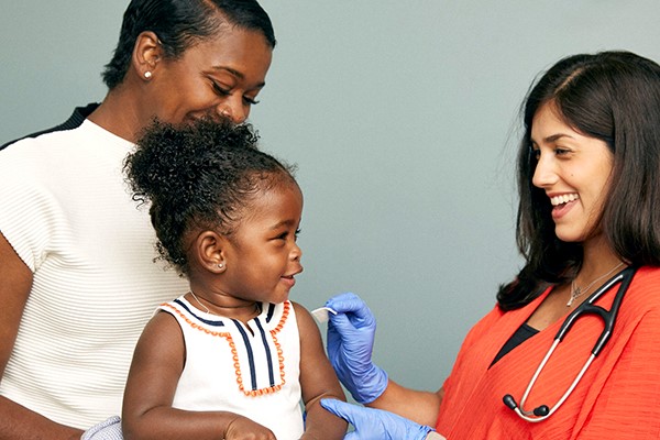 A toddler sits on her mother's lap, as a nurse swabs her arm in preparation for giving a vaccination injection.