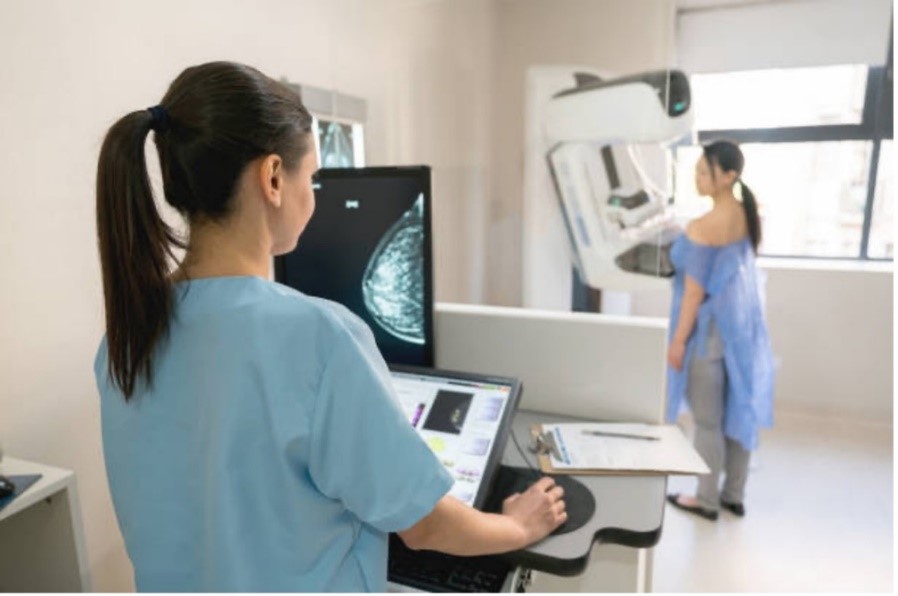 In the foreground, a mammographer views a breast screening image on a computer monitor, while in the background, a woman is having a breast screening in a mammogram machine.